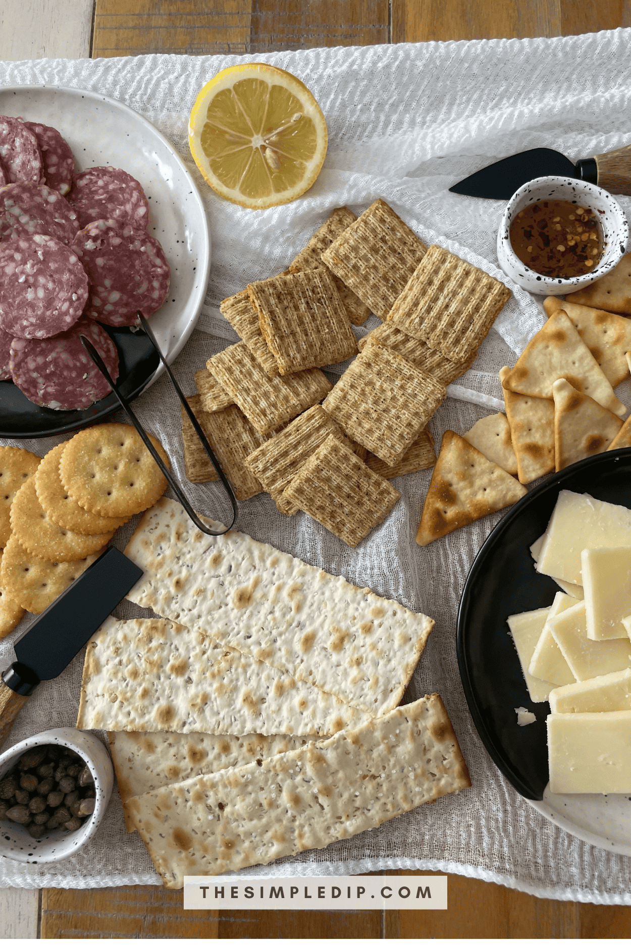 This is an image of a beautiful cracker and charcuterie spread, featuring 4 different kinds of crackers, sliced cheese, sliced soppressata, hot honey, capers, and a sliced half lemon.