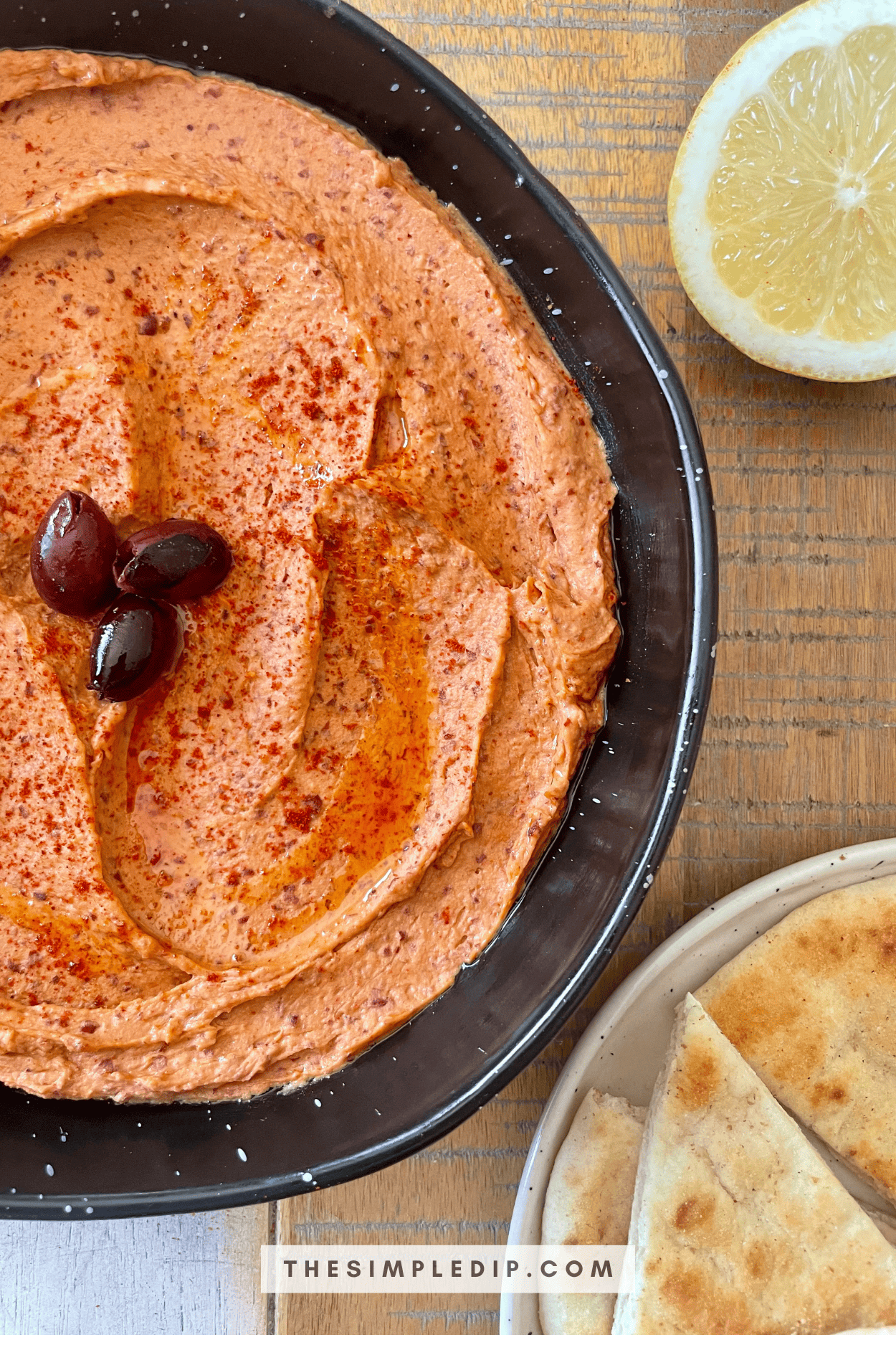 This is a picture of Red Bean Hummus - it has a vibrant orange color with a dusting of red paprika and a trio of olives in the center to elevate the presentation. It's accompanied by a lemon wedge and a plate of sliced pitas.