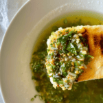 This image depicts an herby, nutty roughly-chopped pesto smothered in golden olive oil to make the perfect Pesto Dipping Oil. A warm, grilled slice of bread is the vehicle for this dip.